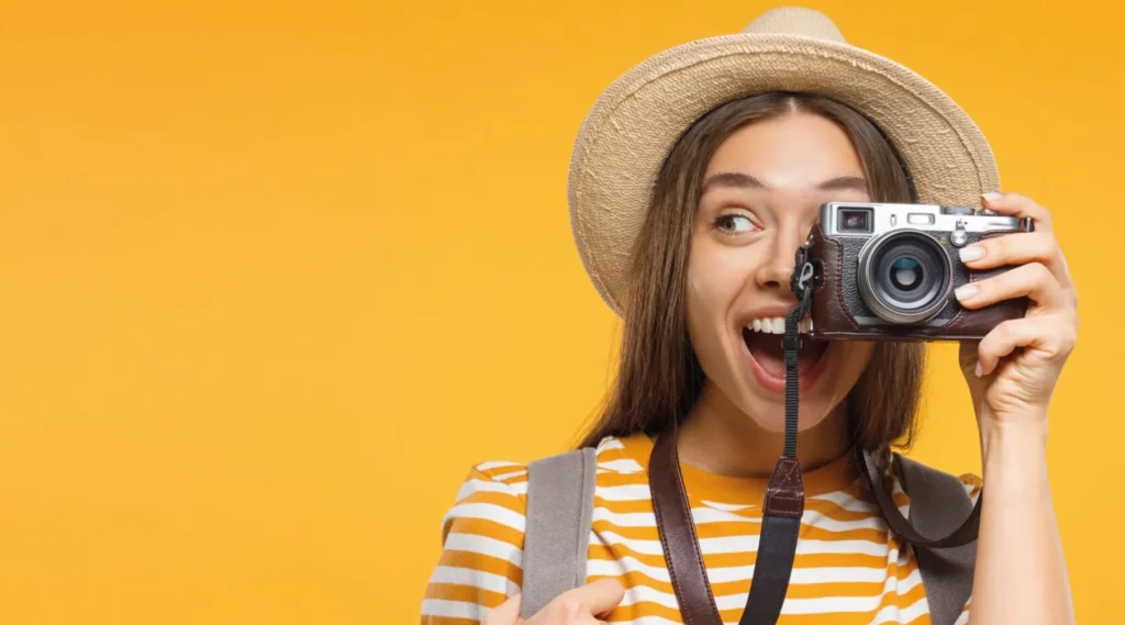 Nell'immagine c'è una ragazza che scatta una fotografia con una vecchia macchina fotografica. Lei indossa un cappello di paglia, una tshirt a righe bianche e gialle e sorride. Lo sfondo è giallo.