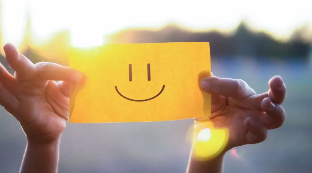 Nell'immagine ci sono le mani di un bambino che reggono un foglio giallo sul quale è stato disegnato uno smile nero