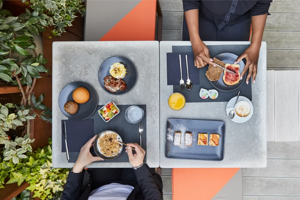 Nell'immagine si vedono due persone dall'alto che fanno colazione. La persona a destra spalma della marmellata su una fetta di pane. Di fronte ha delle marmellatine, un bicchiere di succo, un cappuccino e un piatto con 4 fettine di torta. La persona a sinistra sta mischiando del muesli e dello yogurt e di fronte ha un bicchiere d'acqua, una macedonia, due panini e un piatto con bacone uova.