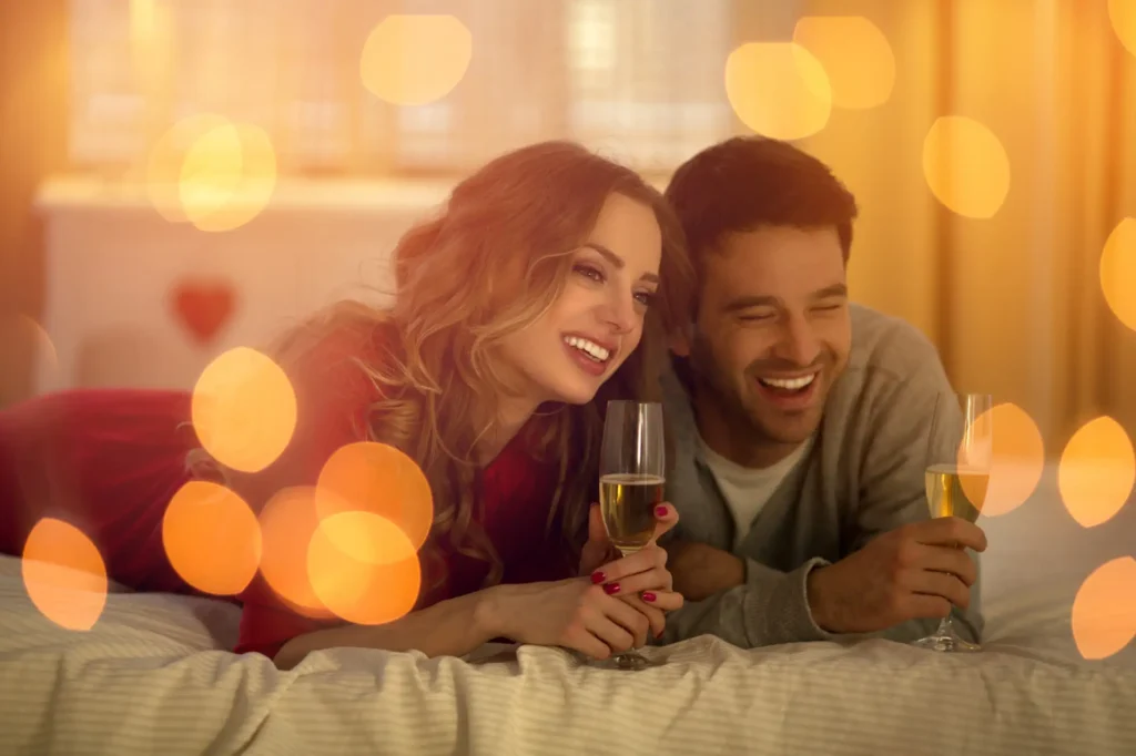 Nell'immagine ci sono una coppia formata da un ragazzo e una ragazza che sono sdraiati pronti sui gomito su un letto. Entrambi tengono tra le mani un bicchiere di spumante e sorridono.