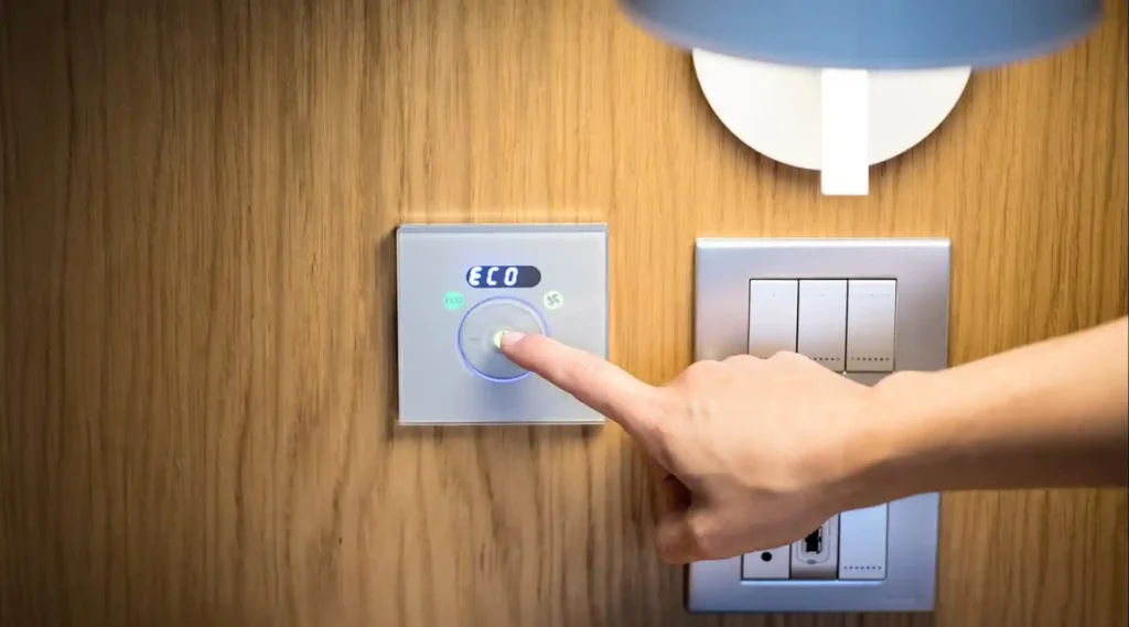 Nell'immagine si vede il dito di una persona che regola un termostato digitale impostandolo su eco. Accanto al termostato ci sono degli interruttori e sopra una lampada