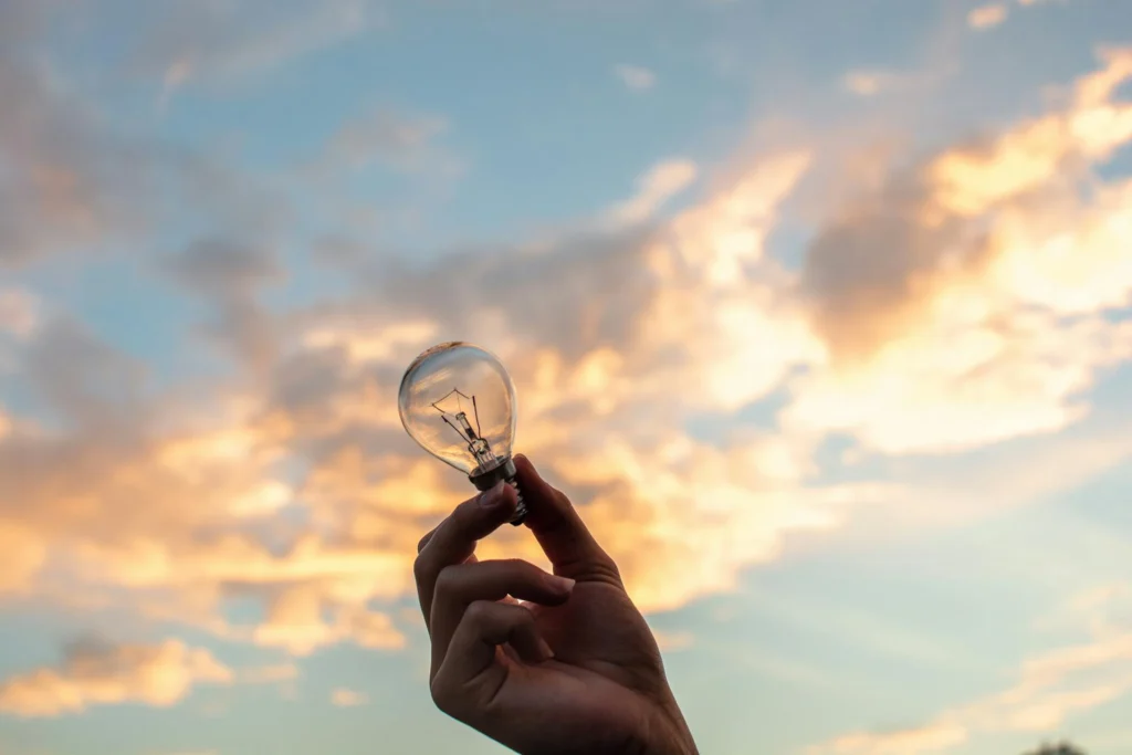 Una mano tiene tra le dita una lampadina a incandescenza con sullo sfondo un cielo al tramonto con delle nuvole che diventano arancioni